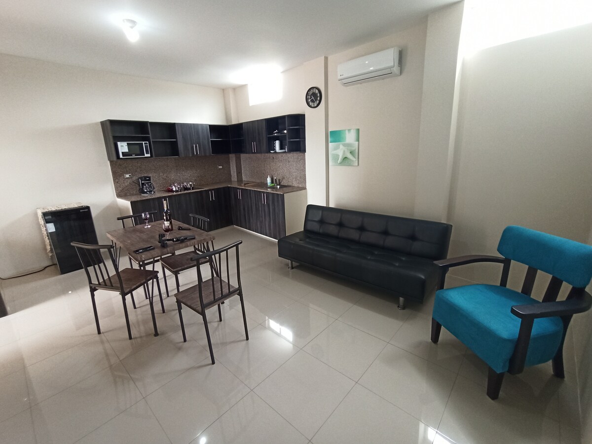 An open living space features a sleek black sofa and an accent chair in blue. A wooden dining table with four chairs is positioned near the modern kitchen, complemented by granite countertops. Bright lighting enhances the airy atmosphere.