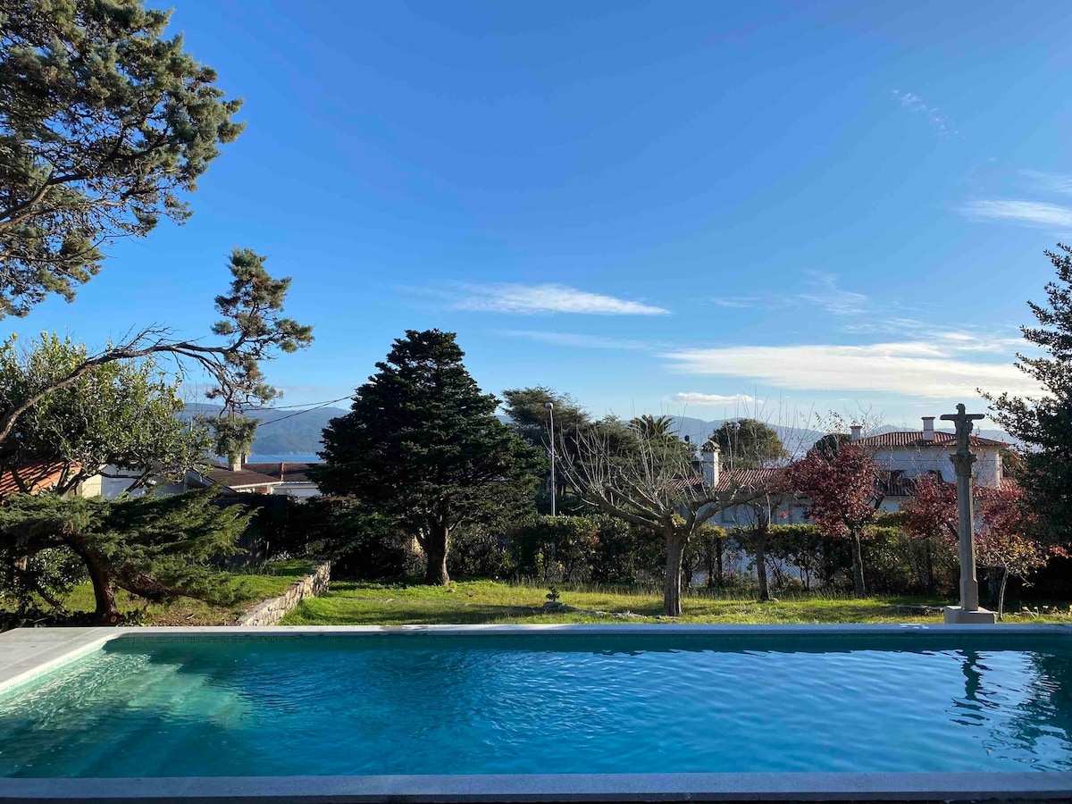A serene pool is set within a spacious outdoor area, surrounded by lush greenery and trees. The clear blue sky provides a bright backdrop, while distant hills add depth to the landscape. The inviting pool invites relaxation and enjoyment in the open air.