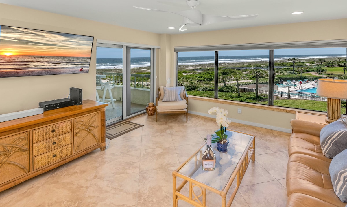 The living area features a neutral palette with a comfortable seating arrangement. A large television is mounted on the wall, while expansive windows offer views of the ocean and pools beyond. Natural light fills the space, complementing the elegant decor.