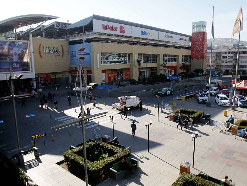 A modern shopping mall exterior is shown with a spacious plaza in front. Visitors are seen strolling, with some sitting on benches. Various storefronts are visible, along with green landscaping and light poles. The bustling atmosphere of the area is highlighted by nearby vehicles and pedestrians.
