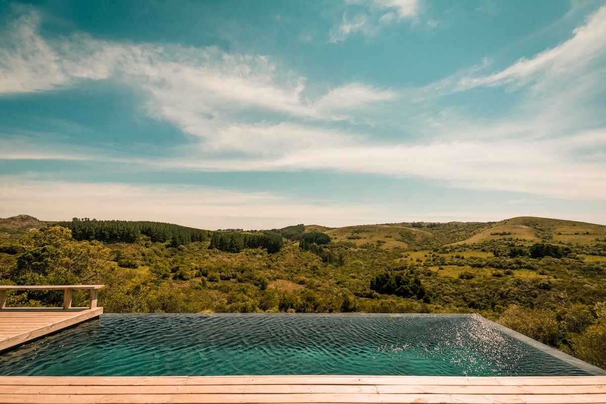 An infinity pool extends across a wooden deck, offering unobstructed views of rolling green hills and a clear sky. Lush vegetation surrounds the area, enhancing the sense of tranquility and connection with nature.