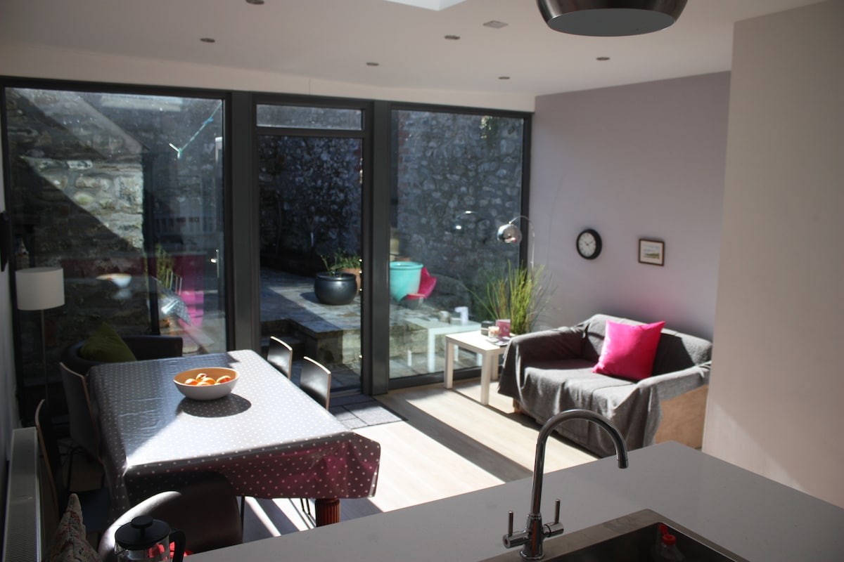 The living area features a combination of natural light from large glass doors and cozy furnishings. A seating area with a gray sofa and colorful cushions invites relaxation. A dining table, equipped for meals, is positioned nearby, with a bowl of oranges as a centerpiece.