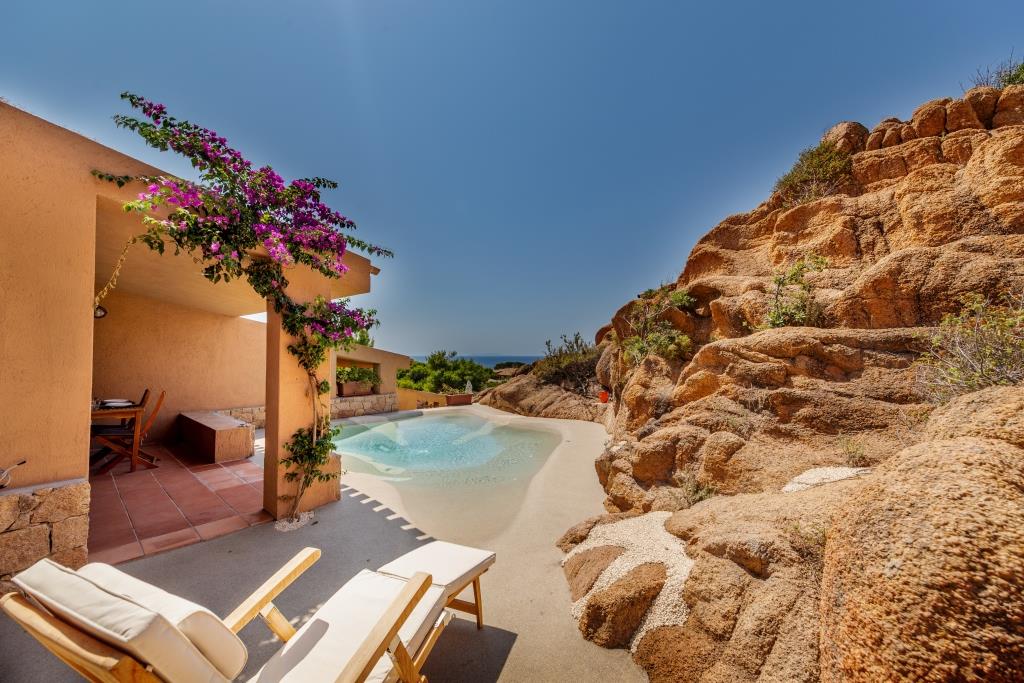 A natural hydromassage pool is framed by rugged rocks and colorful vegetation. Sun loungers are positioned nearby, offering a view of the sea. The entrance to a living area is visible, with a relaxed patio space inviting leisure under the clear blue sky.