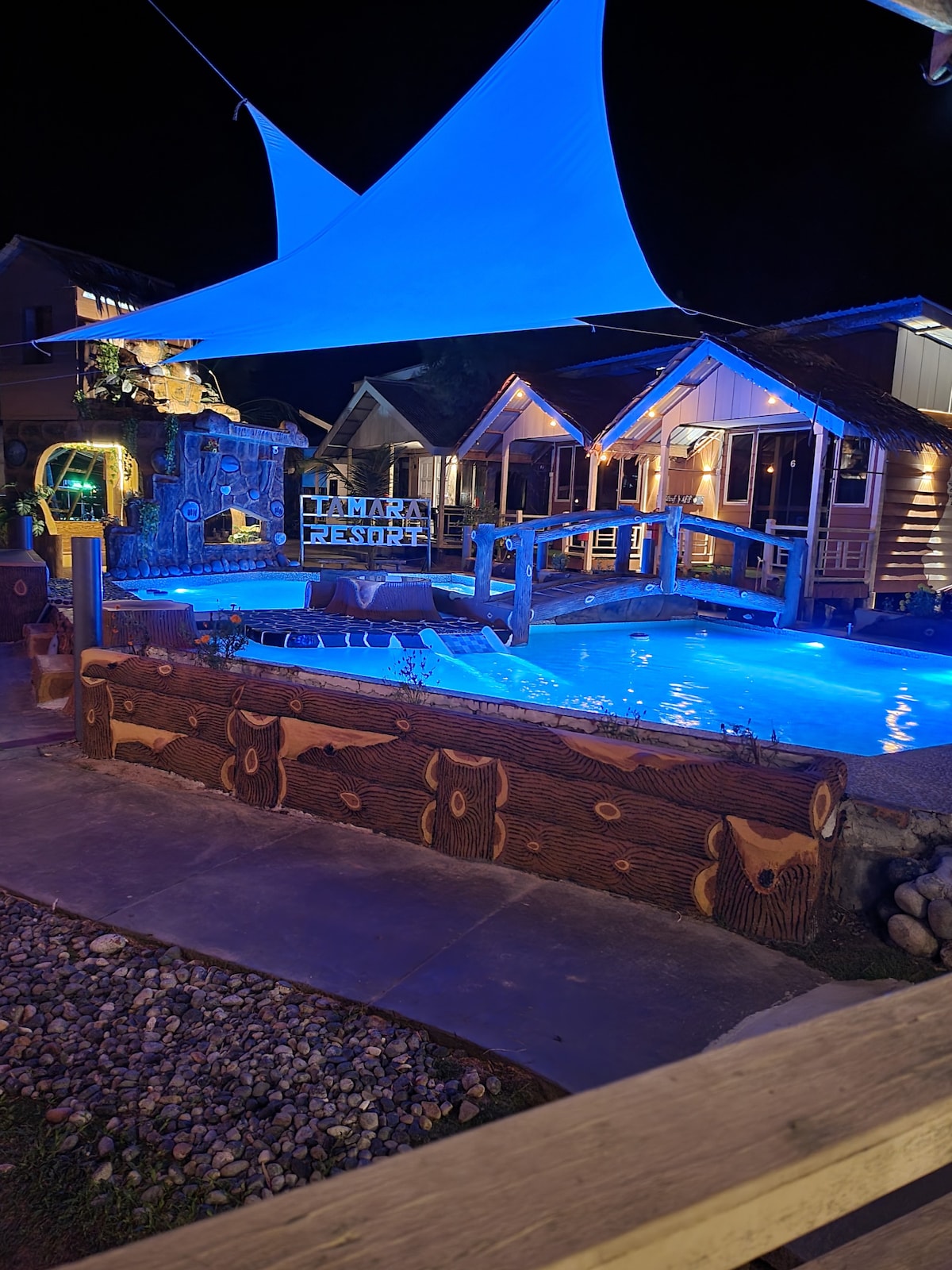 A tranquil outdoor area is illuminated in blue light, showcasing a swimming pool and surrounding deck. The pool is framed by wooden logs, with cozy cabins visible nearby. A large canopy provides shade, enhancing the inviting atmosphere of this peaceful retreat.