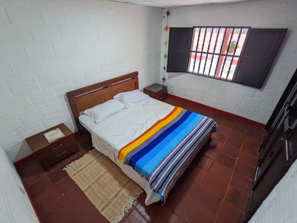 A cozy bedroom is presented with a spacious bed adorned with a multi-striped blanket. Two nightstands are positioned on either side of the bed. Natural light filters through the window, highlighting the smooth, white walls and earthy-toned floor.