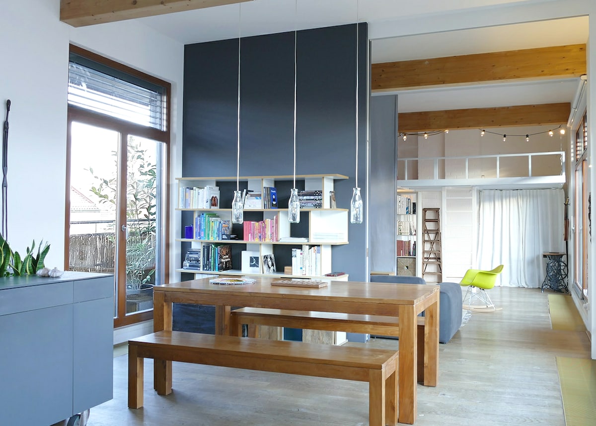 A spacious dining area features a long wooden table paired with matching benches. A minimalist bookshelf filled with books stands against a dark wall. Natural light enters through large windows, illuminating the room with soft brightness.