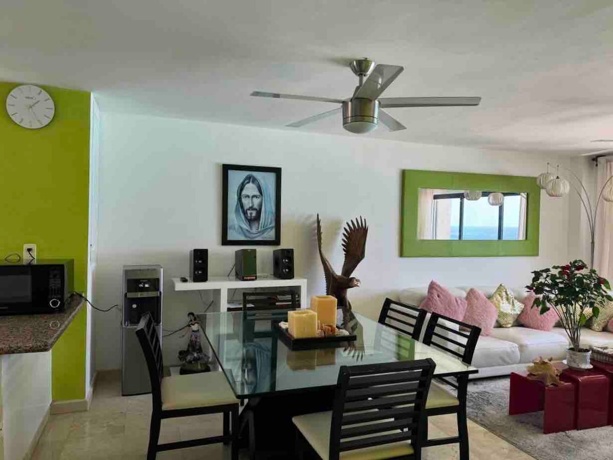The dining area features a glass table surrounded by black chairs, complemented by a vibrant green wall and a decorative mirror. A cozy seating area is visible in the background, with light hues and soft furnishings. Appliances and speakers are neatly arranged, enhancing the modern aesthetic.