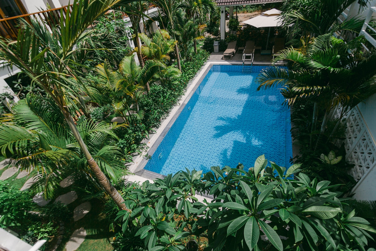 A clear swimming pool is surrounded by lush tropical foliage, creating a vibrant green frame. Lounge chairs are positioned strategically along the pool's edge, inviting relaxation. Sunlight reflects gently on the water's surface, highlighting the serene ambiance of the space.