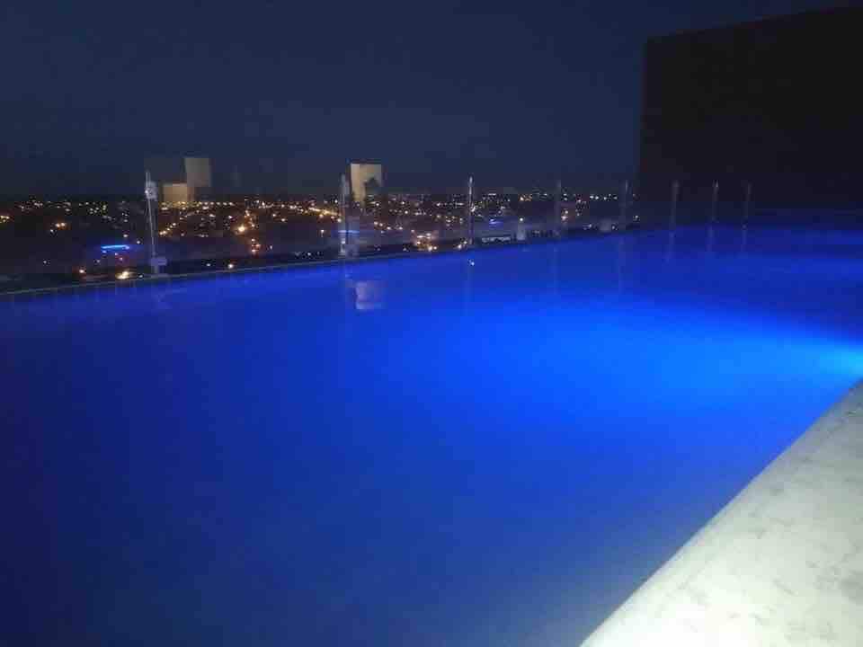 A rooftop pool is seen illuminated in soft blue light against a nighttime cityscape. The surrounding area showcases the distant glow of city lights, providing a serene atmosphere. The pool's edge is lined with transparent fencing for safety and unobstructed views.