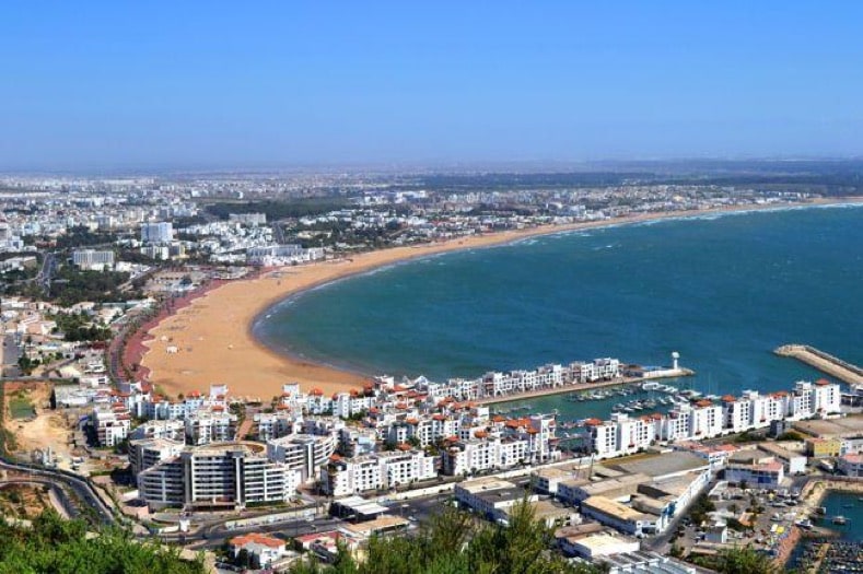 A panoramic view showcases the coastline of Agadir, featuring a long expanse of sandy beach alongside the ocean. The bustling waterfront is dotted with boats, while modern buildings line the shore. The scene is complemented by clear blue skies and distant mountains framing the landscape.