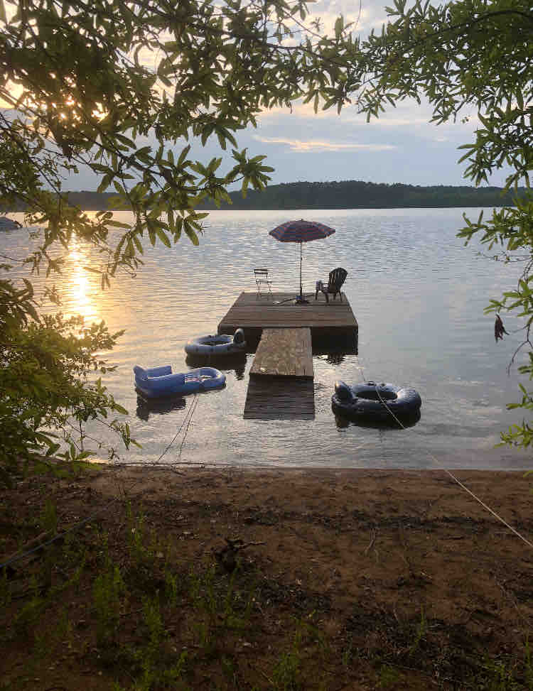 A wooden dock extends into a calm lake, with a single chair shaded by an umbrella. Two inflatable lounges float nearby, surrounded by water reflecting the soft glow of the setting sun. Trees frame the scene, adding to the tranquil atmosphere.