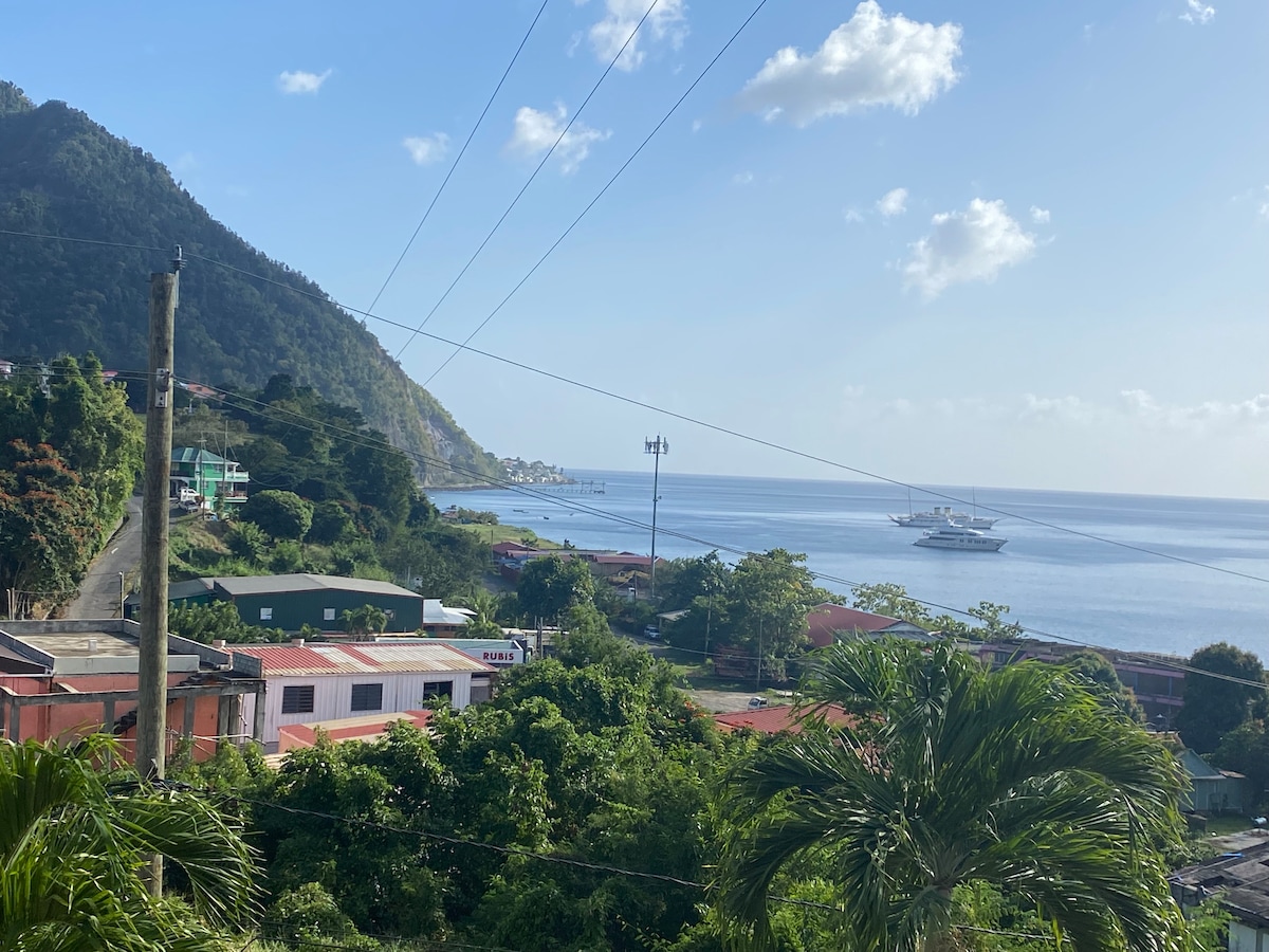 A scenic view reveals a coastal landscape with lush greenery in the foreground. The blue waters of the ocean extend toward the horizon, framed by hills in the background. Soft clouds drift in a clear sky, while a boat is anchored nearby.