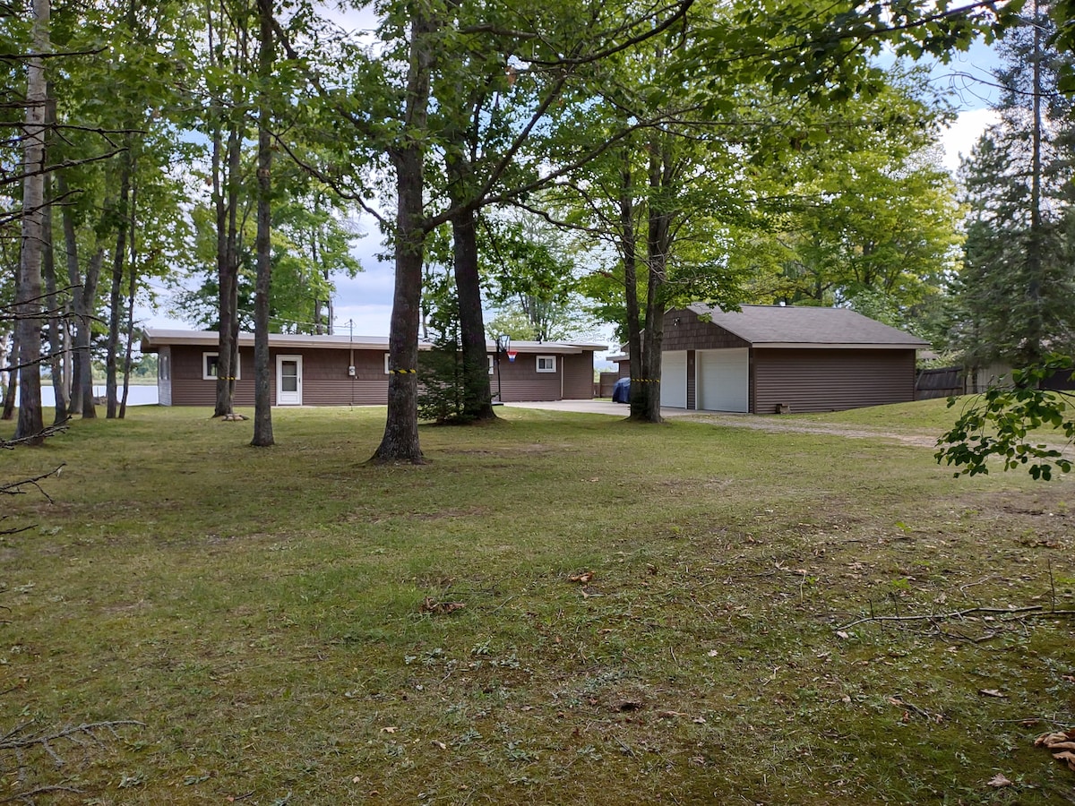 The cabin, set amidst lush greenery, is surrounded by towering trees. A spacious lawn provides ample outdoor space. The structure features large windows, while a detached garage is visible on the right. The serene lake can be glimpsed through the trees in the background.