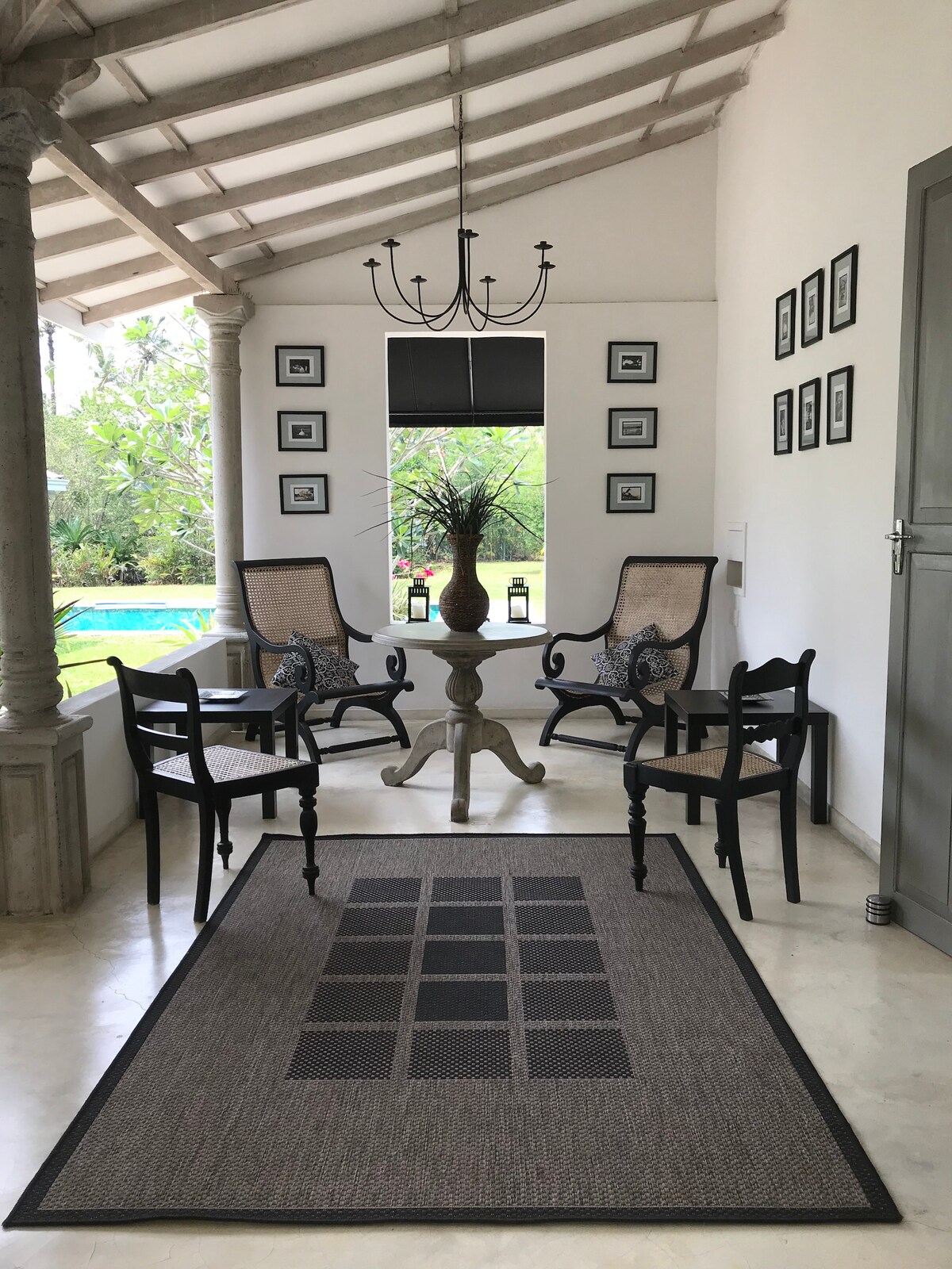 A serene sitting area features two elegant armchairs and two sleek side chairs arranged around a round table. A decorative vase rests on the table, while framed photographs adorn the walls. Natural light brightens the space, highlighting the view of the lush garden outside.