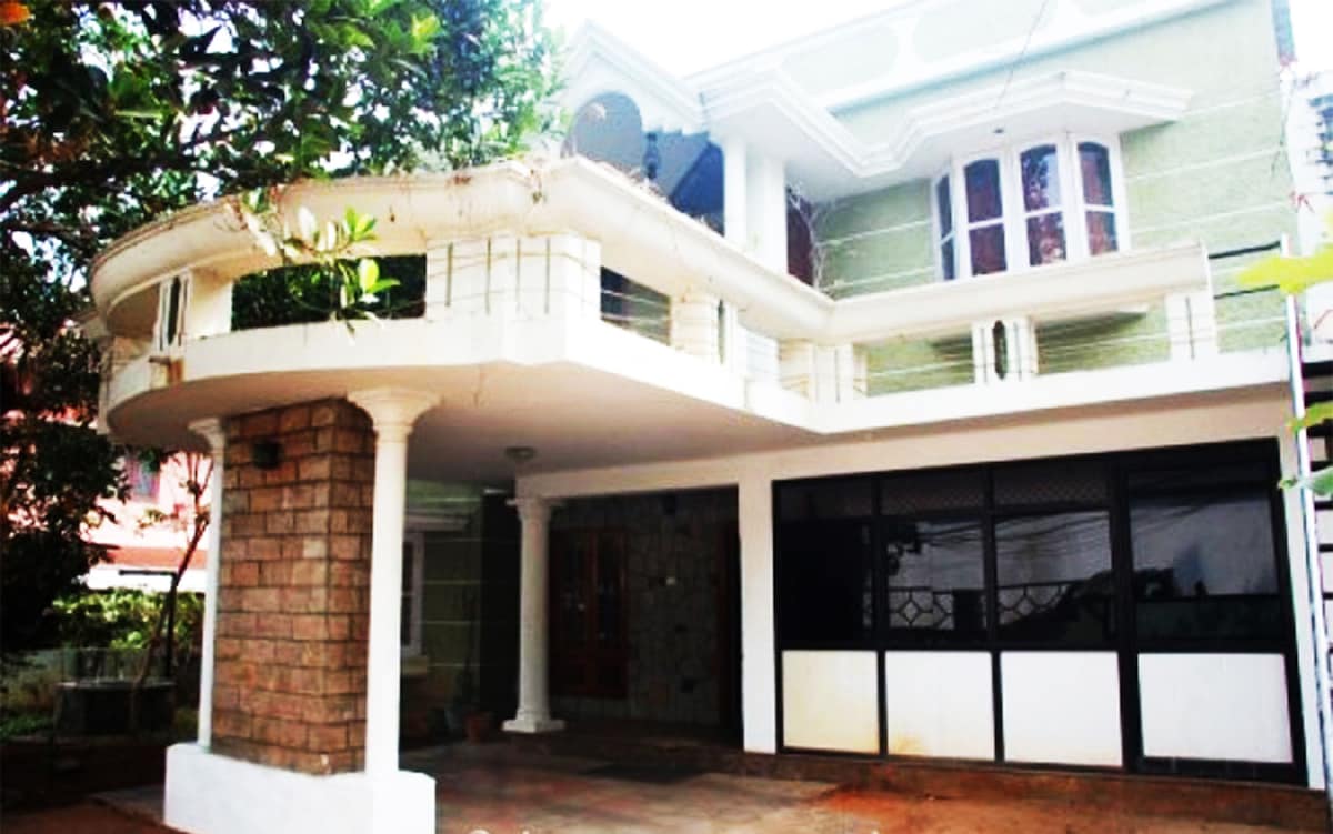 The villa exhibits a grand facade with a mix of stone and painted surfaces. Curved balconies are supported by elegant columns, while large windows provide ample natural light. A spacious entrance leads to a comfortable outdoor area surrounded by greenery.
