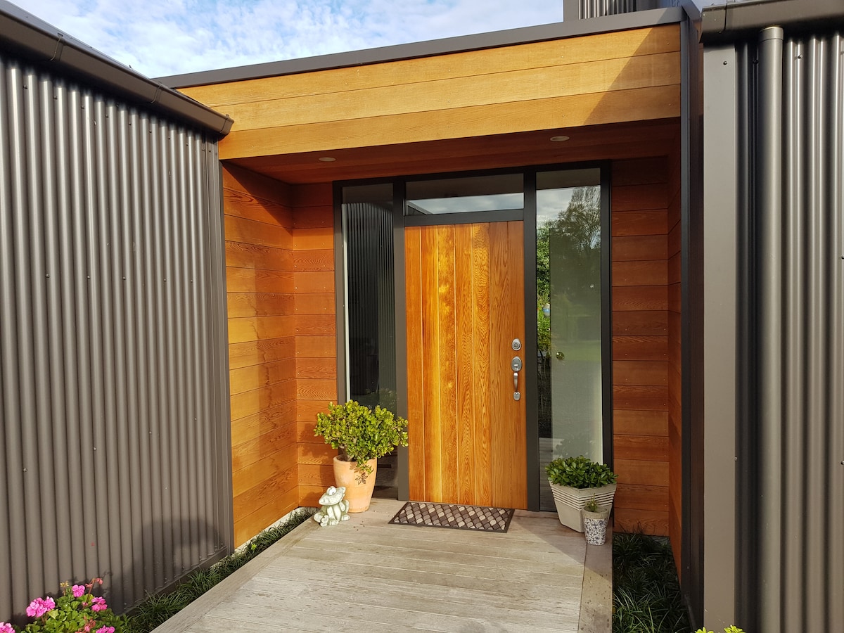 The entrance of the house features a wooden front door set within an overhanging awning, framed by contemporary metal siding. Potted plants are placed on either side of the entryway, and a welcome mat is positioned in front of the door, enhancing the inviting appearance.
