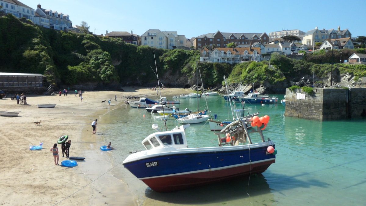 A view of a sandy beach is presented, with several boats anchored in calm waters. People are seen engaging in activities on the shore, while greenery rises in the background, framing the coastal scene. A few buildings are visible on the hillside above the beach.