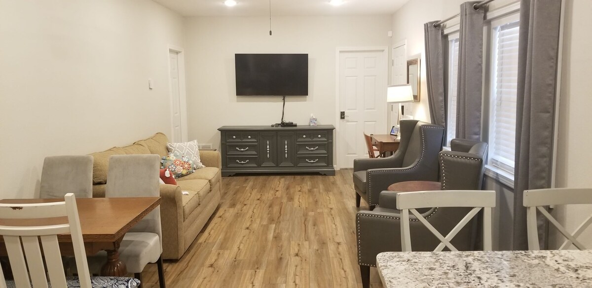 A spacious living area is featured, highlighting a large flat-screen TV mounted on the wall above a grey console. Comfortable seating arrangements include a beige sofa, a grey armchair, and a dining table with chairs. Natural light filters through window coverings.