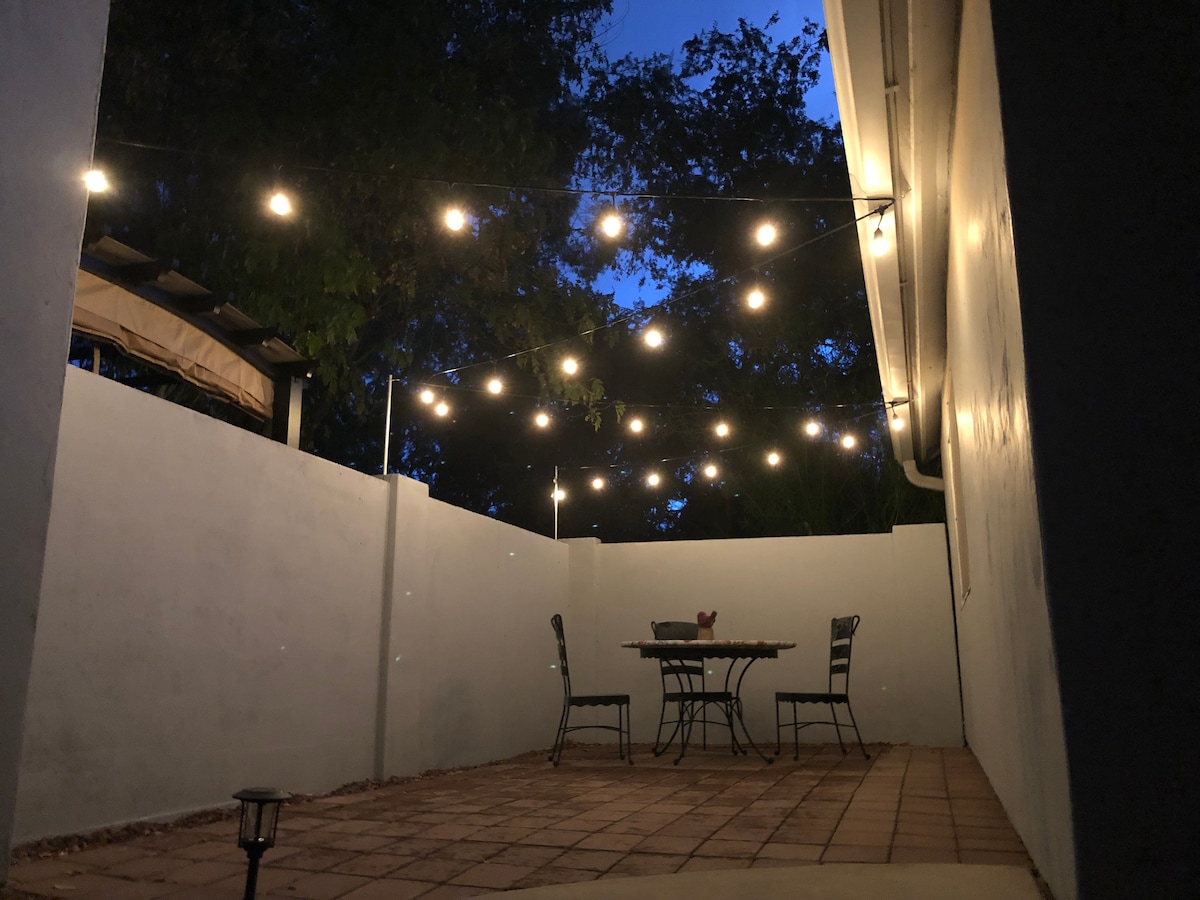A private patio area is shown, featuring a dining table with four chairs beneath softly glowing string lights. The surrounding walls provide a sense of seclusion, while the evening sky adds a tranquil ambiance.