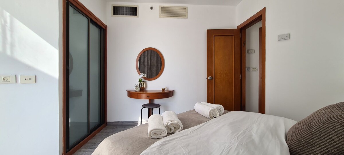 A cozy bedroom is designed with neutral colors. A spacious bed is covered with soft linens, and neatly rolled towels are placed at the foot. A round mirror hangs above a small table, complemented by a flower vase. Natural light enters through a nearby window.