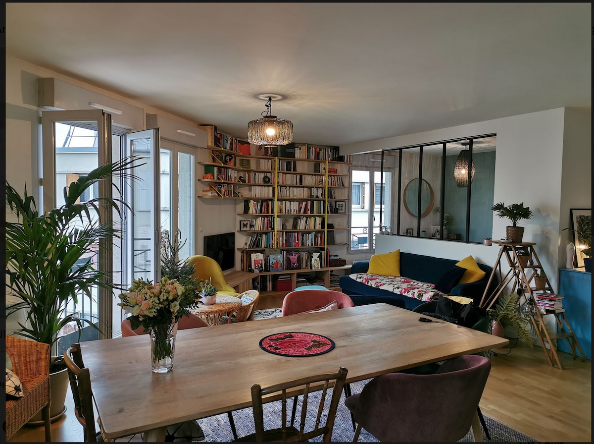 The living area features a large wooden dining table adorned with a decorative centerpiece. A bookshelf filled with books lines the wall, while comfortable seating is positioned nearby. Large windows allow natural light in, complemented by decorative plants adding a touch of greenery.