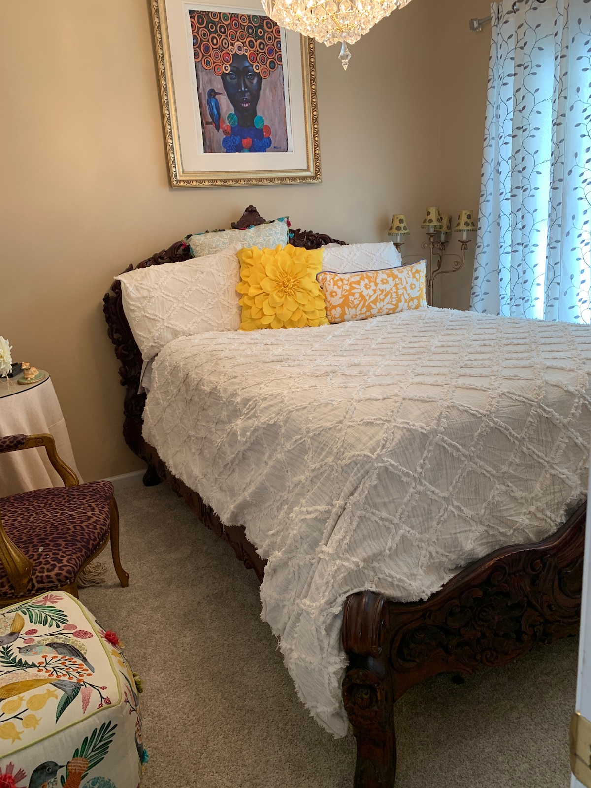 A spacious bed with a textured white coverlet is positioned in a softly lit bedroom. Colorful decorative pillows add a vibrant touch. Intricate wooden furniture can be seen alongside elegant curtains, while a framed artwork enhances the inviting atmosphere.