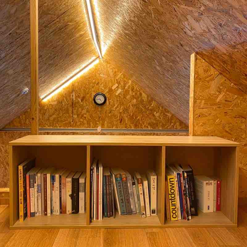 A wooden bookshelf is displayed in a lofted space, filled with a variety of books. The warm tones of the wood complement the natural textures of the sloped ceiling, accented by soft lighting. A round clock is mounted on the wall, adding a functional element to the cozy design.