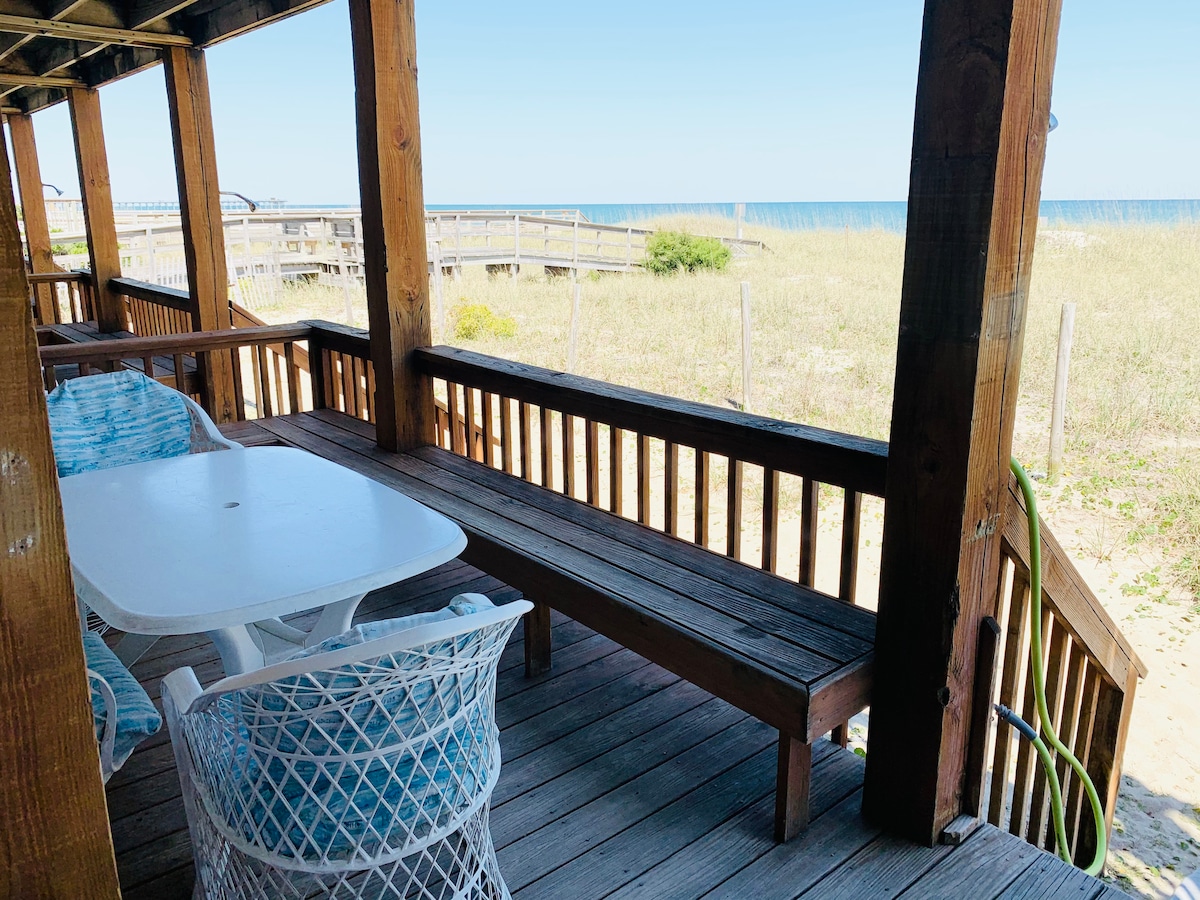 An ocean-side porch is depicted, featuring a long wooden bench and a white table suitable for dining. Two white wicker chairs complement the setting, which overlooks sandy beach grass and stretches towards the ocean. Sunlight brightens the area, enhancing the inviting outdoor space.