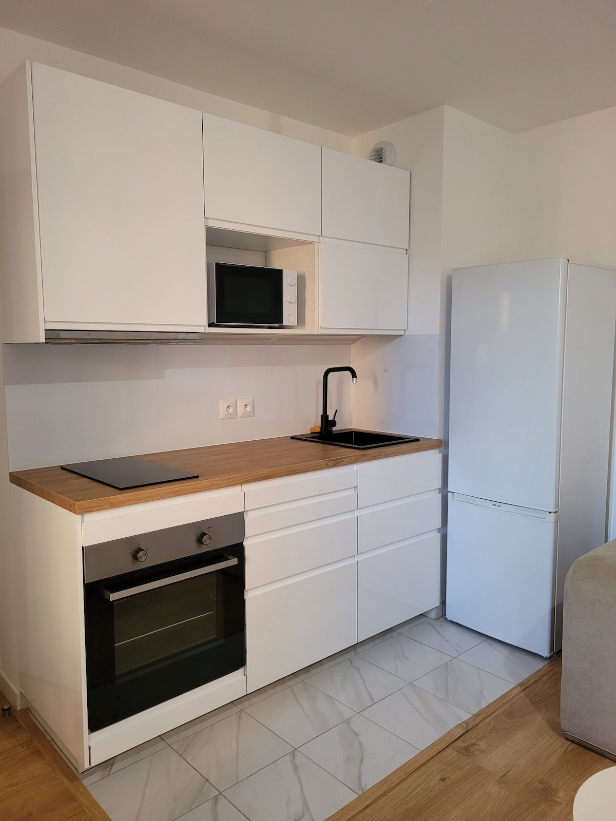 A modern kitchen layout is presented, featuring sleek white cabinets and a contrasting wooden countertop. Essential appliances such as a microwave and oven are included, along with a refrigerator. A black sink with a matching faucet is installed, enhancing the kitchen's functionality.