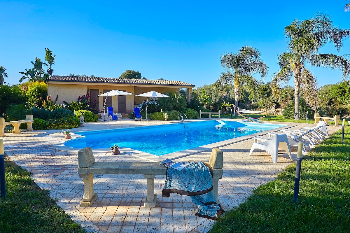 An outdoor swimming pool is surrounded by palm trees and lush greenery. Loungers and umbrellas create inviting relaxation areas. A hammock is partially visible, adding a tranquil touch. The pool area is paved with tiles, complementing the natural landscape.