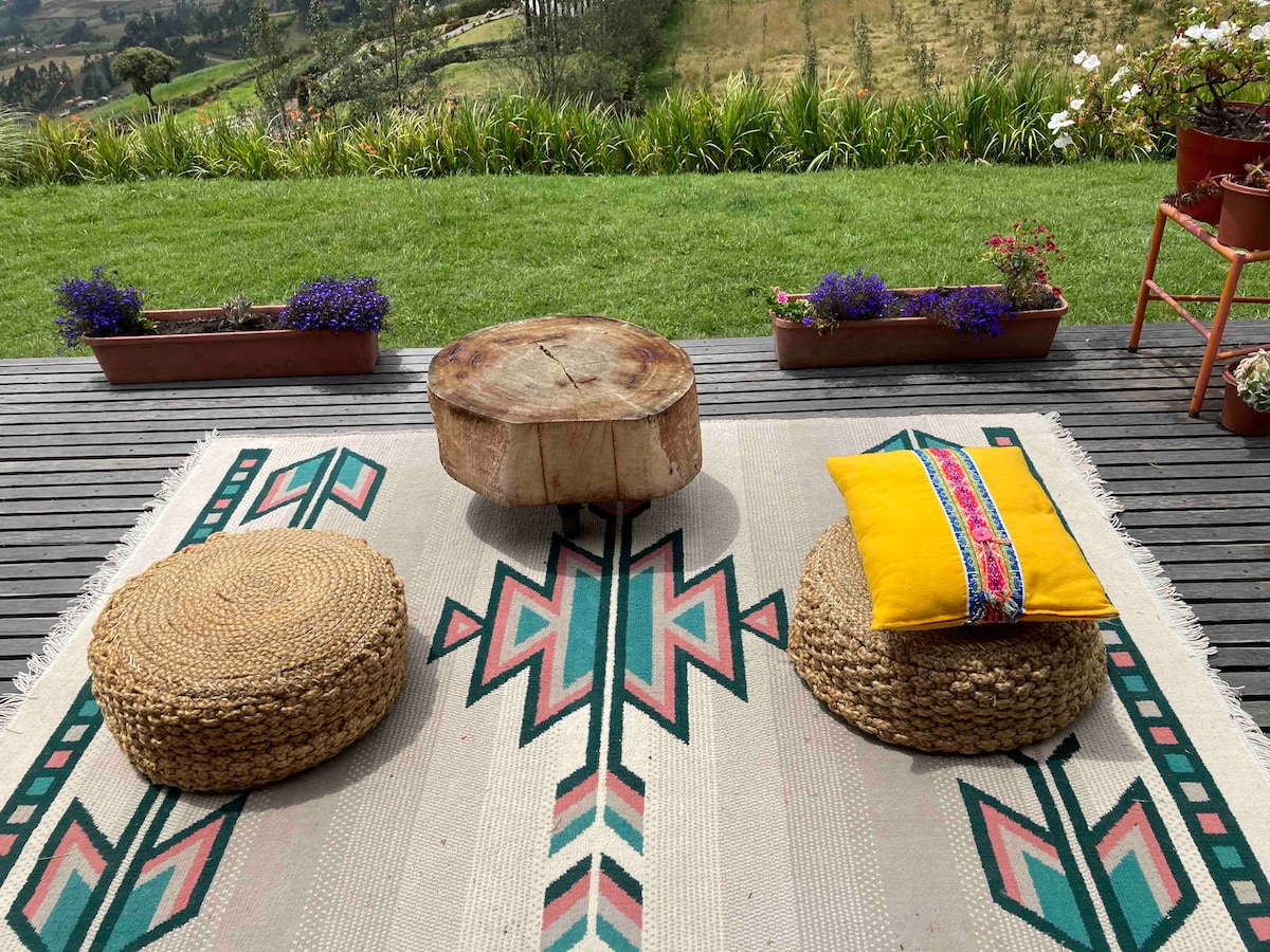 A tranquil outdoor seating area features a rustic wooden table surrounded by woven stools on a vibrant rug. Lush greenery and flower pots are visible in the background, offering a serene view of the surrounding countryside.