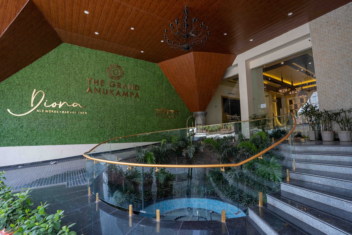 An elegant entrance to The Grand Anukampa features a curved glass railing and a cascading water feature. Lush greenery decorates the walls, complementing the modern architectural design. A stylish chandelier hangs from the ceiling, creating an inviting atmosphere for guests arriving at the building.