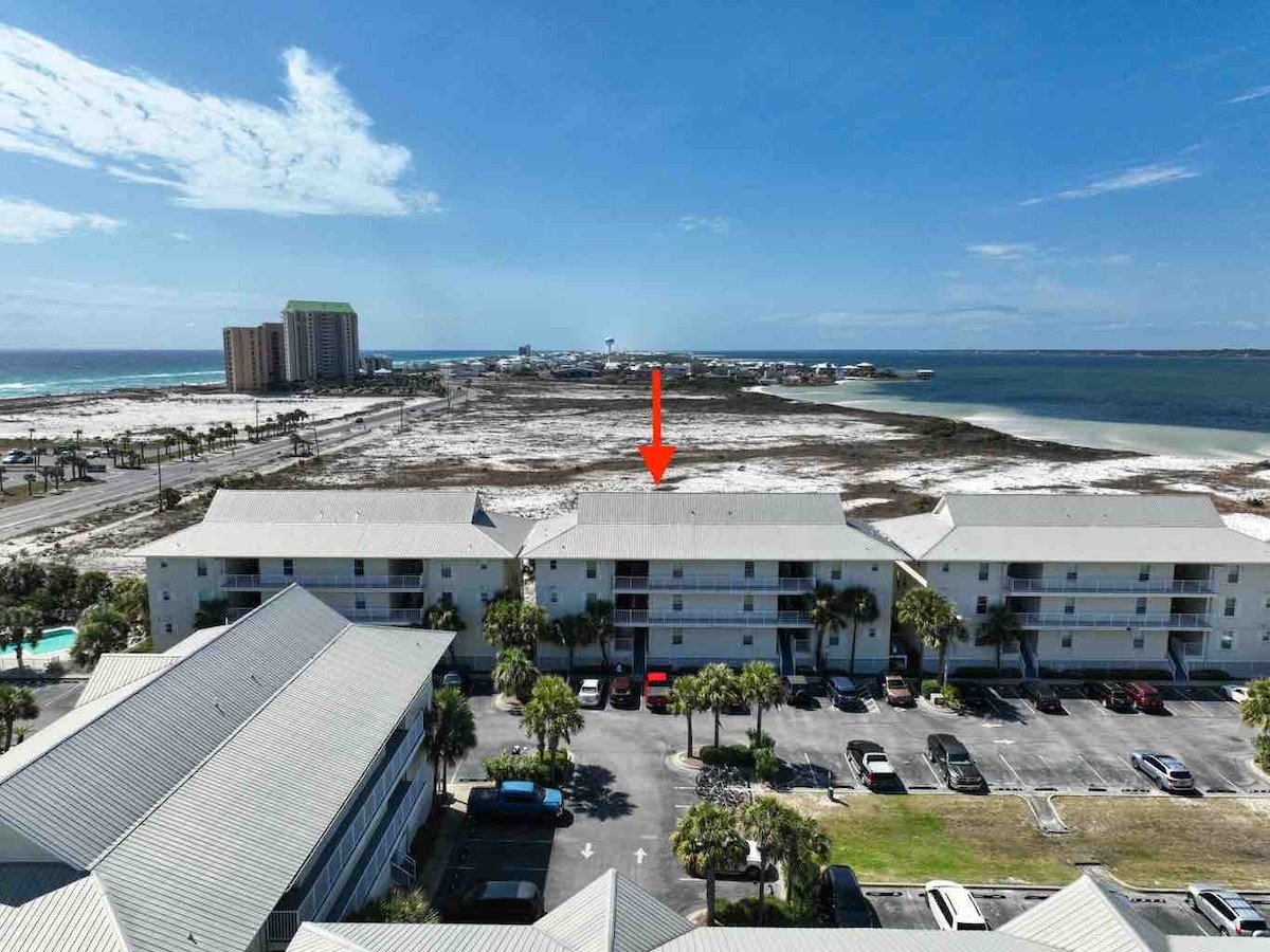 An aerial view captures the beachfront condo complex along with parking spaces and palm trees. The Gulf of Mexico is visible beyond the buildings, showcasing sandy shores and emerald waters. The surrounding area includes a mix of natural landscapes and modern developments.