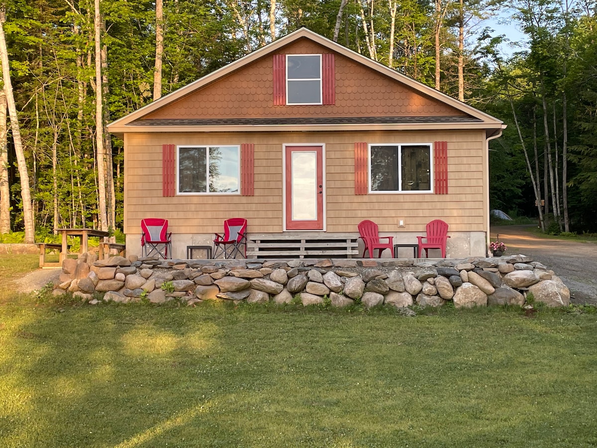 A charming one-story house is surrounded by trees, featuring a welcoming front yard. Four red chairs are positioned on a stone patio, offering a comfortable seating area. The house presents a blend of tan siding with red accents and large windows, providing ample natural light.