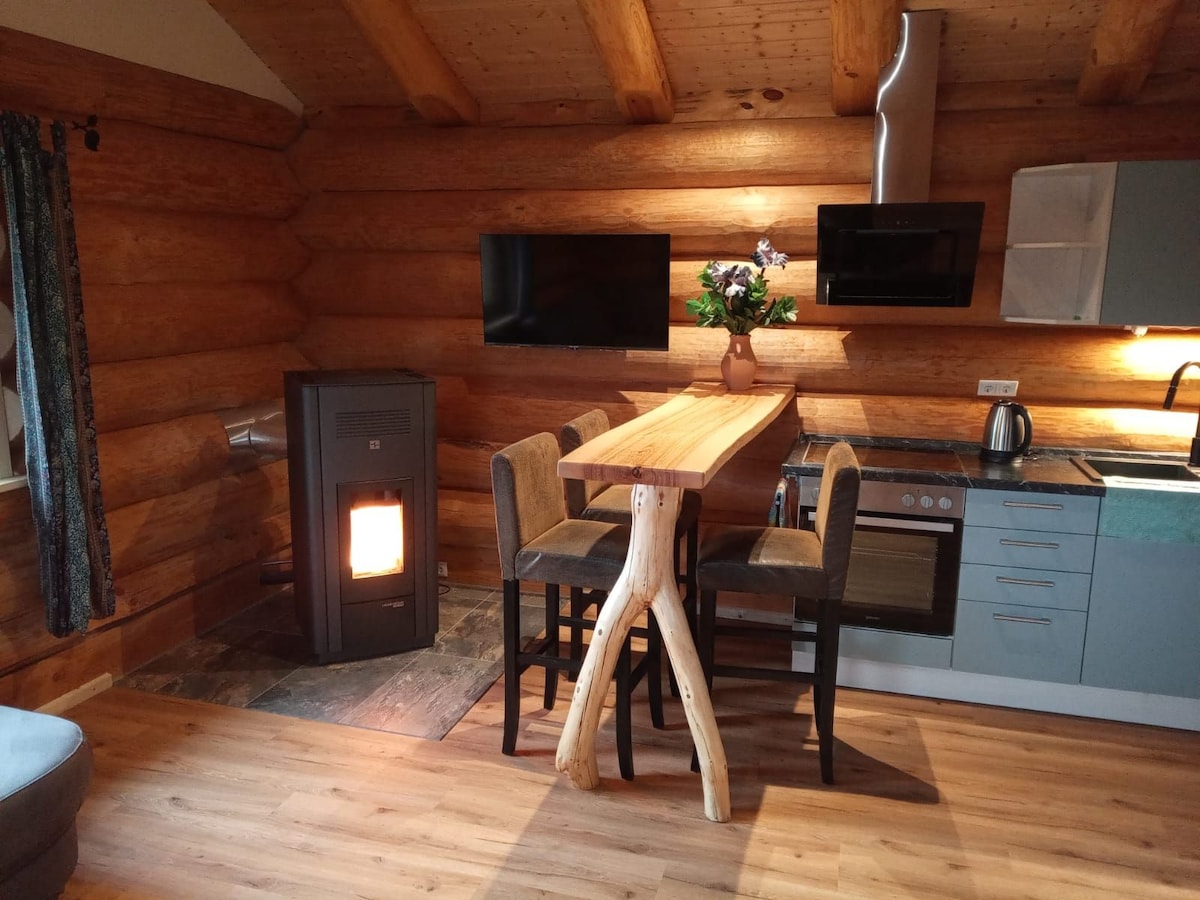 An inviting dining area features a wooden table supported by natural tree branch legs, surrounded by four upholstered chairs. A wood-burning stove adds warmth to the space, while modern kitchen appliances are visible in the background, complementing the rustic log cabin walls.
