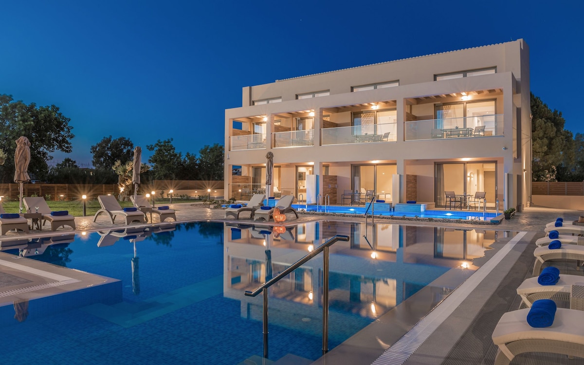 A modern two-story complex is positioned beside a swimming pool. The pool area features lounge chairs arranged around it, with soft lighting reflecting on the water. Balconies are visible on the upper floor, providing outdoor seating options.