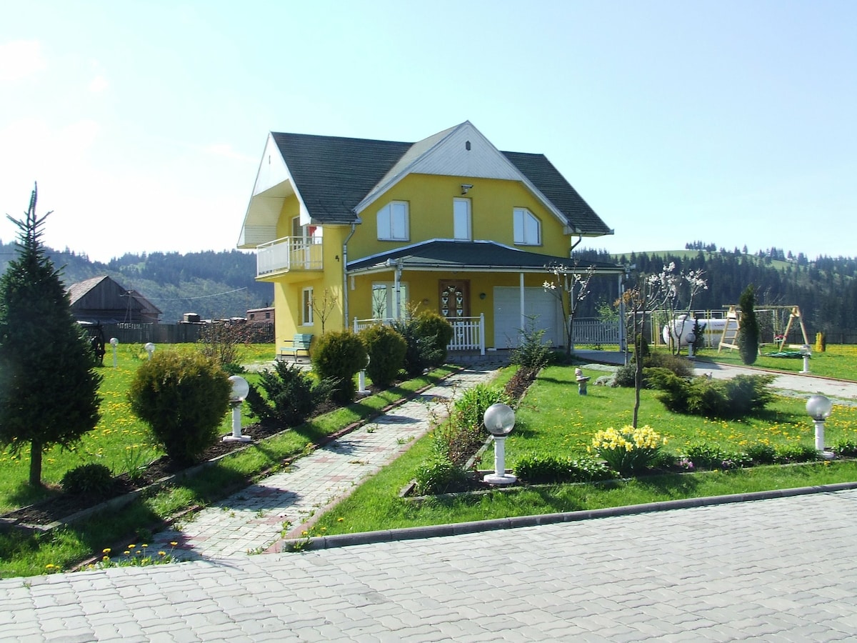 A charming two-story yellow house is situated amidst a landscaped garden featuring manicured shrubs and flowering plants. A stone walkway leads up to the entrance, with a swing set and trees visible in the background under a clear blue sky.