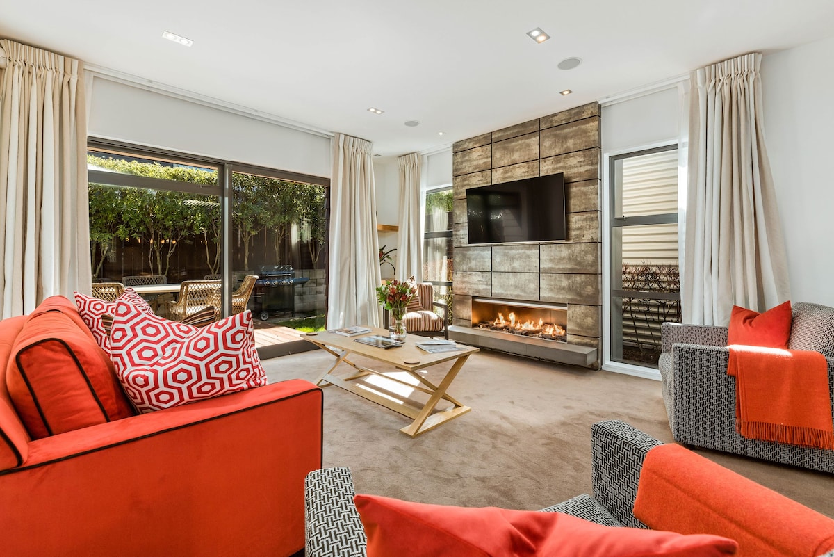 The living area features large glass doors that connect to a private garden, allowing natural light to flood the space. Comfortable sofas, in complementary colors, are positioned near a modern gas fireplace and a wall-mounted Smart TV, creating a relaxed and inviting ambiance.