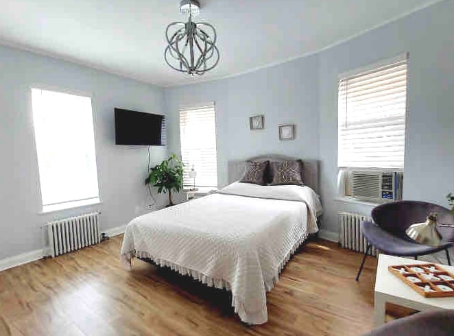 A bright bedroom showcases a spacious bed covered in a delicate white quilt. Large windows allow natural light to fill the room, and a mounted television is visible on the wall. A decorative plant adds a touch of greenery to the space.