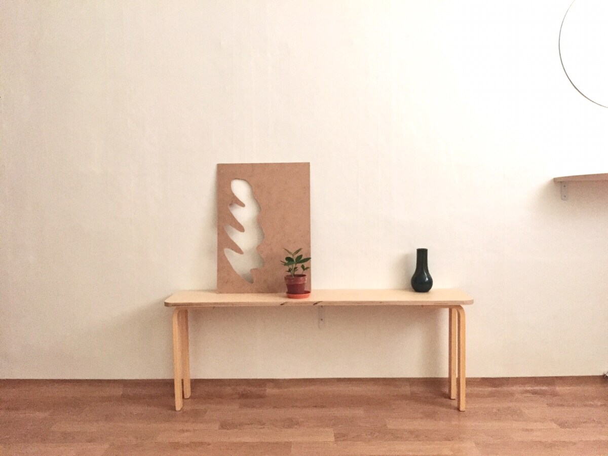 A minimalist wooden bench is positioned against a white wall, featuring a carved art piece resting on it. A small potted plant and a dark vase are placed on the bench, contributing to the serene setting. The warm wooden floor provides a subtle contrast.