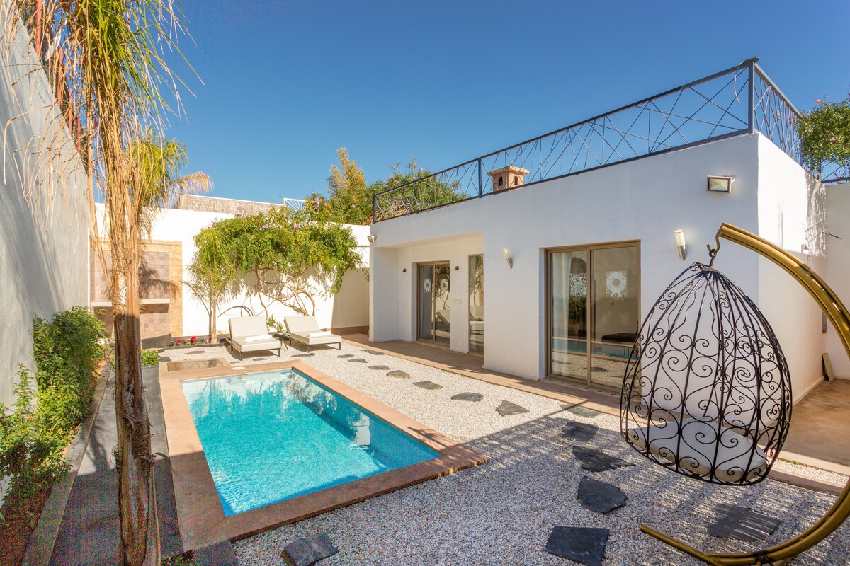 A private pool is surrounded by a landscaped area featuring sun loungers. The villa's exterior is highlighted by large glass doors that open to a bright living space. A hanging chair is positioned nearby, enhancing the relaxing outdoor atmosphere.