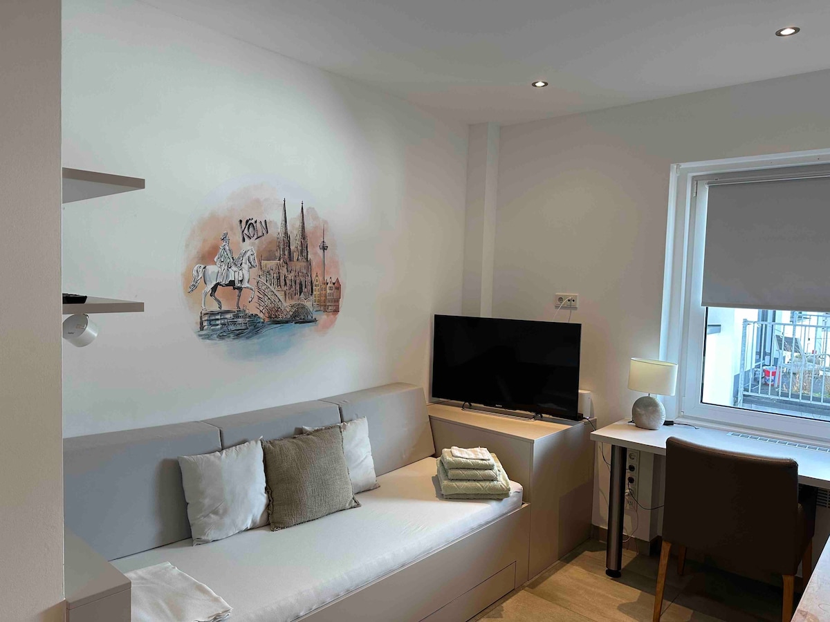 A compact living space features a comfortable sofa bed and a wall-mounted TV. A minimalistic desk is positioned near a window, providing natural light. A mural depicting a cityscape adds a unique touch to the room, while neatly folded towels are positioned on the sofa.