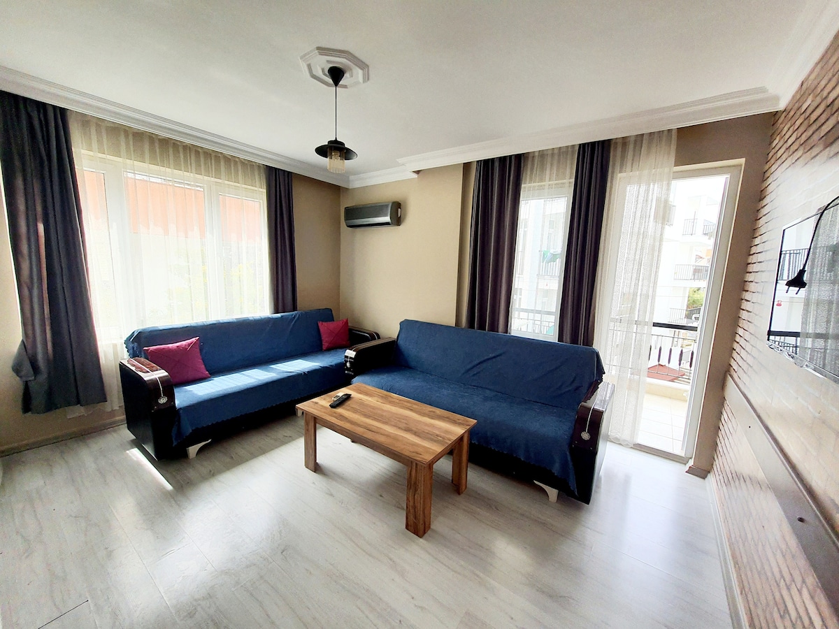 A comfortable living area is presented with two blue sofas, accented by pink cushions. A wooden coffee table sits centrally, and a flat-screen TV is mounted on the wall. Natural light filters through large windows, adorned with light drapes.