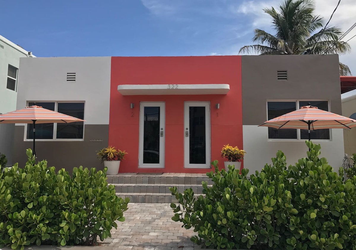 A colorful exterior view of the twin units showcases a modern architectural design. The vibrant red and neutral tones are contrasted by lush greenery and planted pots. Two umbrellas provide shade over small patio areas, inviting relaxation. A paved pathway leads to the entrances.