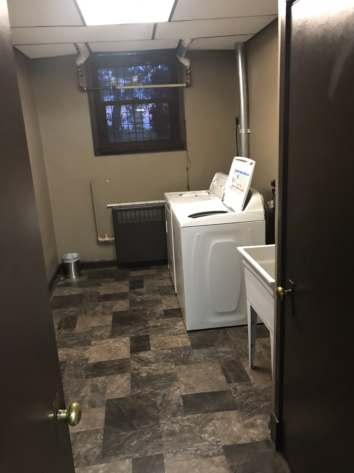 A functional laundry room is equipped with a washing machine and a dryer, positioned against a wall. A utility sink is located beside the machines. Natural light enters through a window, which is slightly obscured by blinds. The floor features dark patterned tiles.
