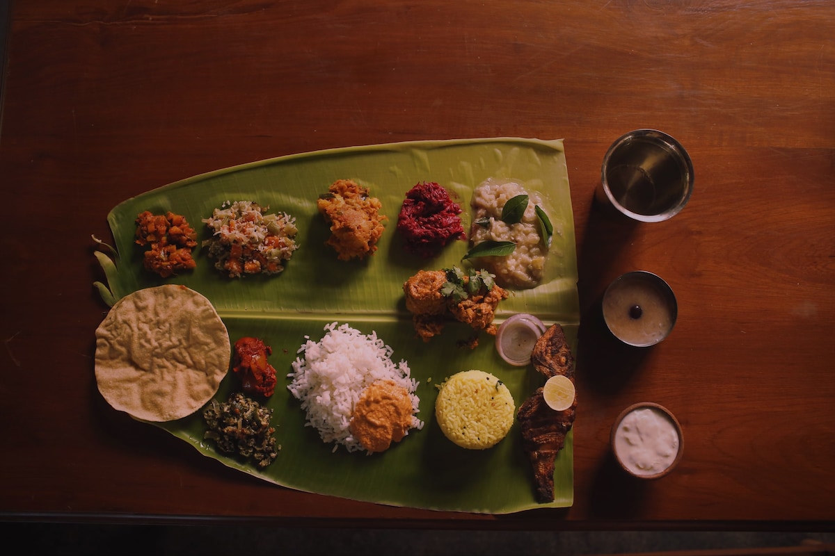 A traditional South Indian meal is arranged on a banana leaf, featuring an assortment of dishes including rice, lentils, vegetables, pickles, and accompaniments. Metal tumblers filled with beverages are placed alongside, providing a complete dining experience in a rustic setting.