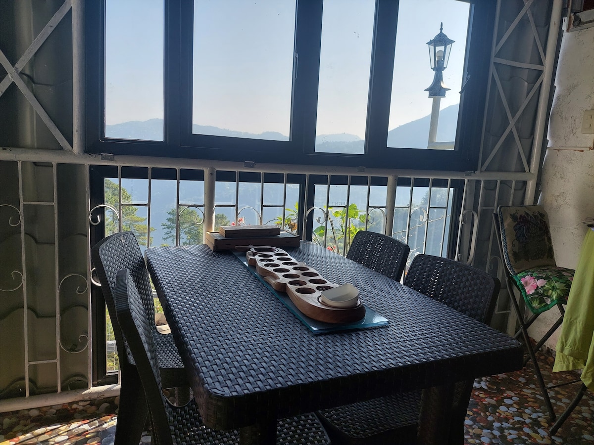 A dining table with dark wicker furniture is positioned near large windows, offering views of distant mountains and greenery. A wooden tray with tea cups is centered on the table, while a decorative lamp stands outside the window, enhancing the serene setting.