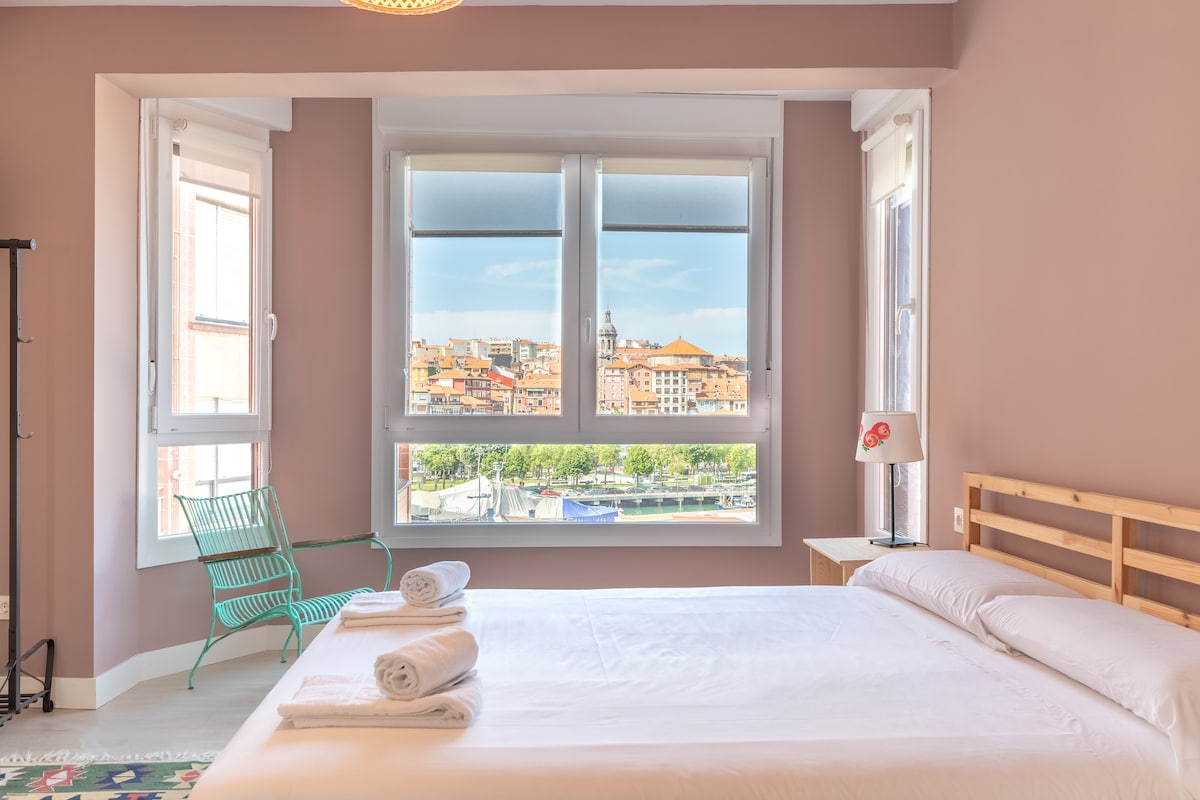 A spacious bedroom features a large bed with fresh linens positioned near wide windows, offering views of the vibrant town. The room is accented by light walls and a stylish bedside lamp, complemented by a green chair in the corner, inviting relaxation.