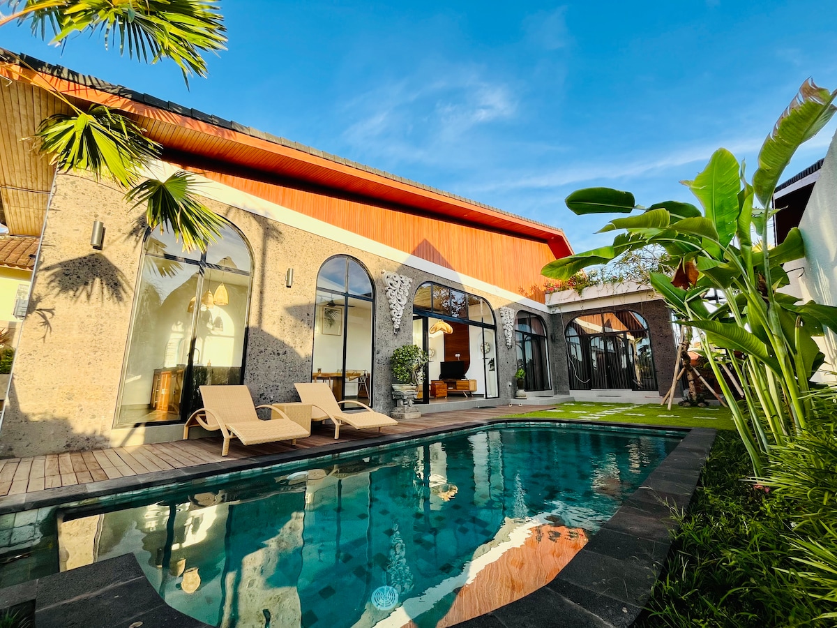 A modern villa is set against a clear blue sky, featuring a tranquil pool surrounded by lush greenery. The exterior showcases large arched windows and a combination of textured stone and vibrant roofing, while sun loungers line the pool area, inviting relaxation.
