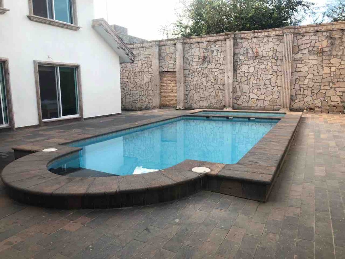 A rectangular pool is surrounded by a stone patio, featuring a smooth edge and steps for entry. The walls behind the pool are constructed of large, light-colored stones, contributing to a sense of privacy. Large windows in the background reflect the natural light.